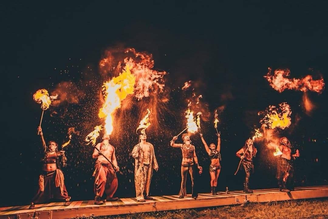 Seven fire performers on an outdoor stage at night, with raised torches and sweeping flames