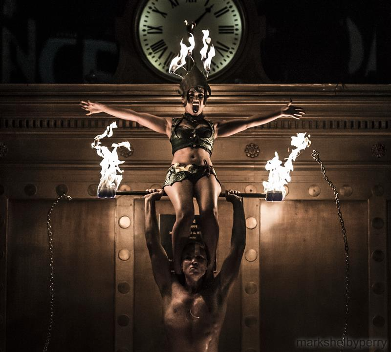 Two performers in an acrobatic fire tableau beneath a clock; photograph by Mark Shelby Perry